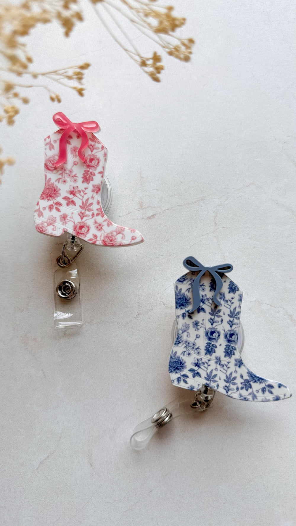 Two floral patterned boot-shaped retractable badge holders with a clear plastic strap. One boot is pink with a pink bow and the other is blue with a blue bow.