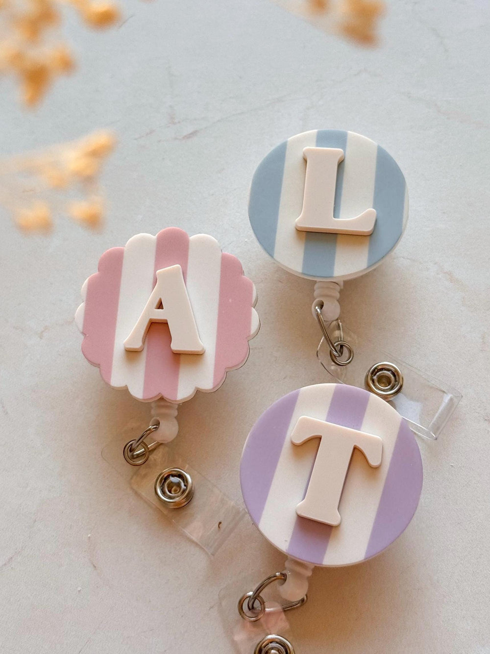 Three personalized badge reels with white, pink, blue, and purple striped backgrounds. Each reel features a different letter: A, L, and T, in a white, raised font. The reels have a clear plastic strap