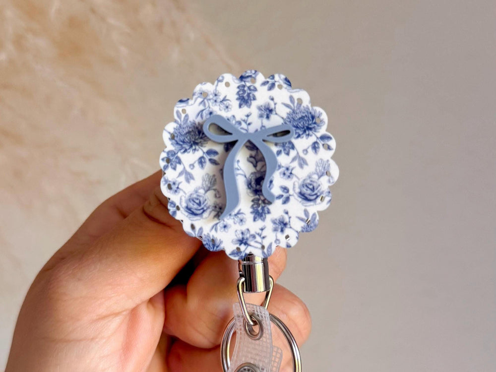 A floral badge reel with a blue bow design. The reel features a white background with blue floral patterns and a scalloped edge. The bow is a solid light blue color.