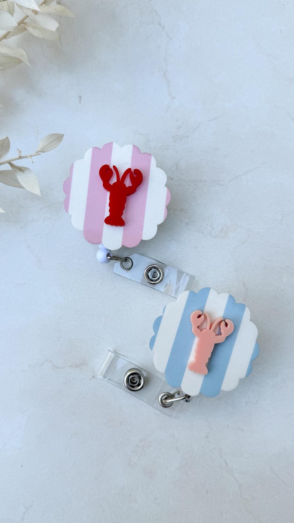 Two badge reels with scalloped edges. One has pink and white stripes with a red lobster, the other has blue and white stripes with a pink lobster. Each reel has a clear plastic clip.