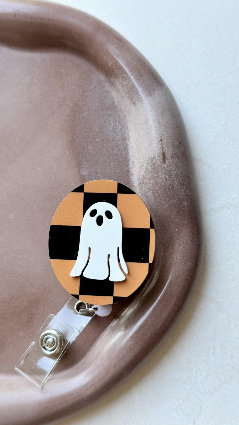 A Halloween-themed badge reel featuring a white ghost design on a black and orange checkered background. The badge reel has a clear plastic strap and a metal clip for attaching to clothing or accessor
