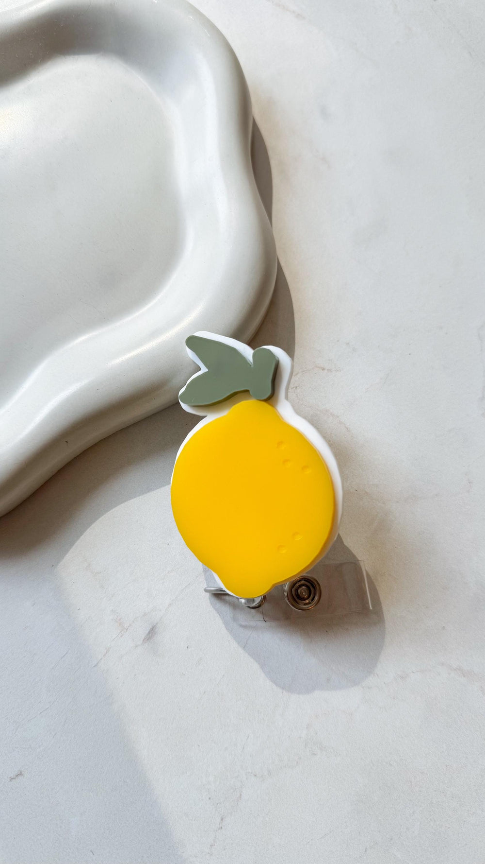 A yellow and white lemon-shaped badge reel with a green leaf accent. The reel has a clear plastic strap and a metal clip. The design is a bright, cheerful illustration.