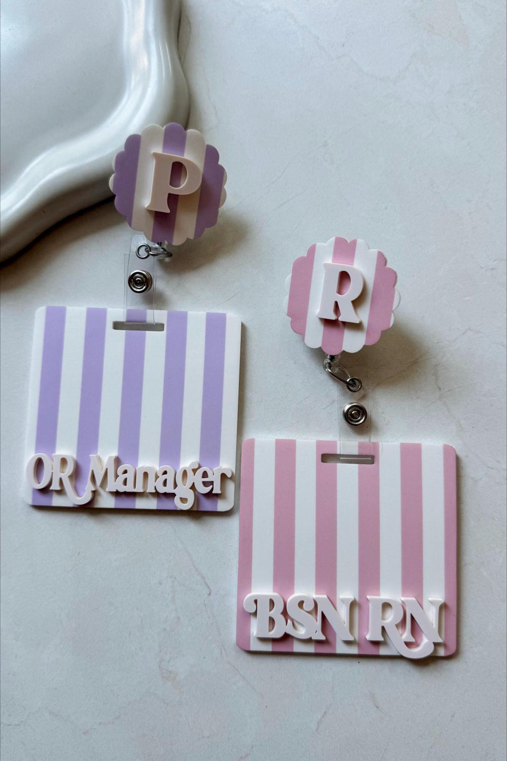 Two badge reels with striped backgrounds and scalloped tops. One is lavender and white with the letter "P" and the text "OR Manager." The other is pink and white with the letter "R" and the text "BSN 
