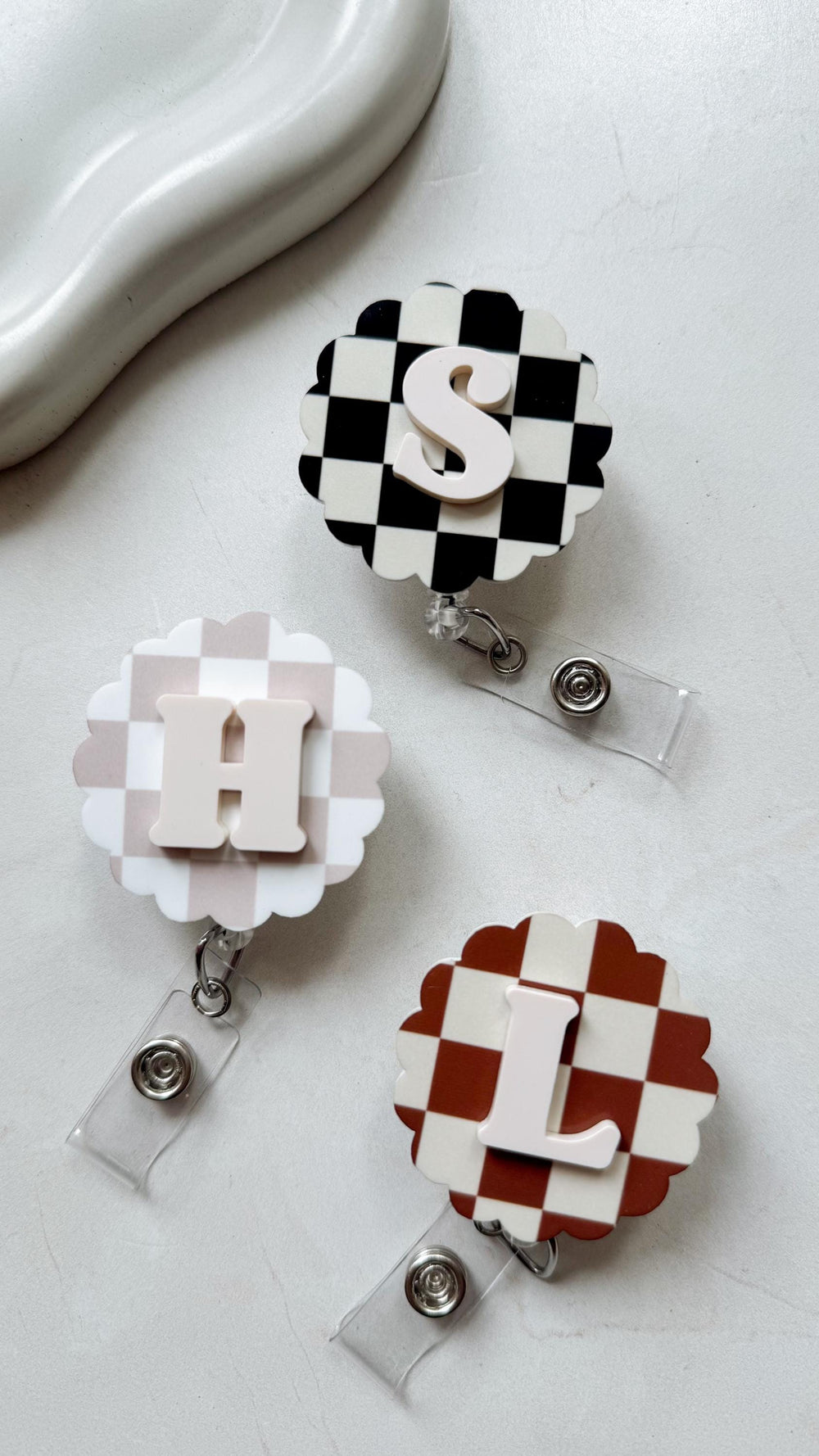 Three badge reels with scalloped edges and checkered patterns. Each reel features a different letter: S in black and white, H in beige and white, and L in brown and white. The letters are in a contras