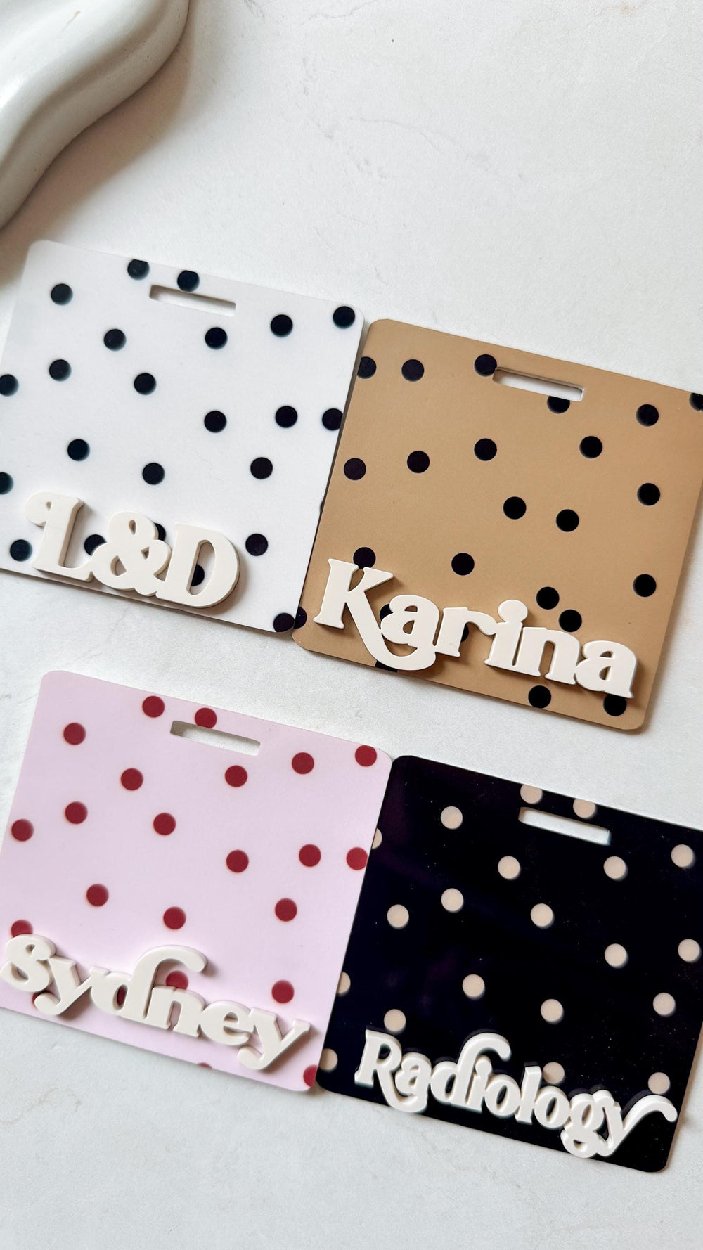 Four name tags with polka dot patterns. The tags are white, tan, pink, and black. The white tag has black dots and the letters "". The tan tag has black dots and the name "Karina". The pink tag has re