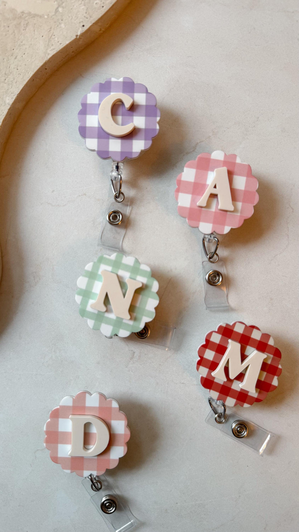 A collection of five badge reels with gingham patterns in various colors: purple, pink, green, peach, and red. Each reel features a different letter: C, A, N, D, and M, in a light beige color.