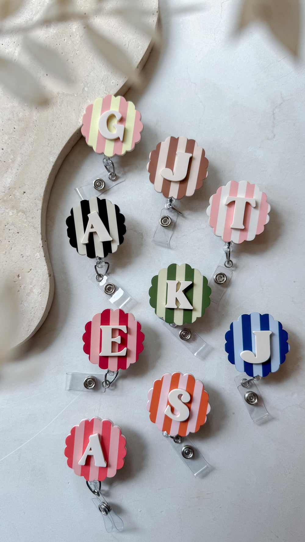 A collection of personalized badge reels with various initial letters. Each reel features a scalloped edge and a striped background in colors like pink, blue, green, and black. The letters are in whit