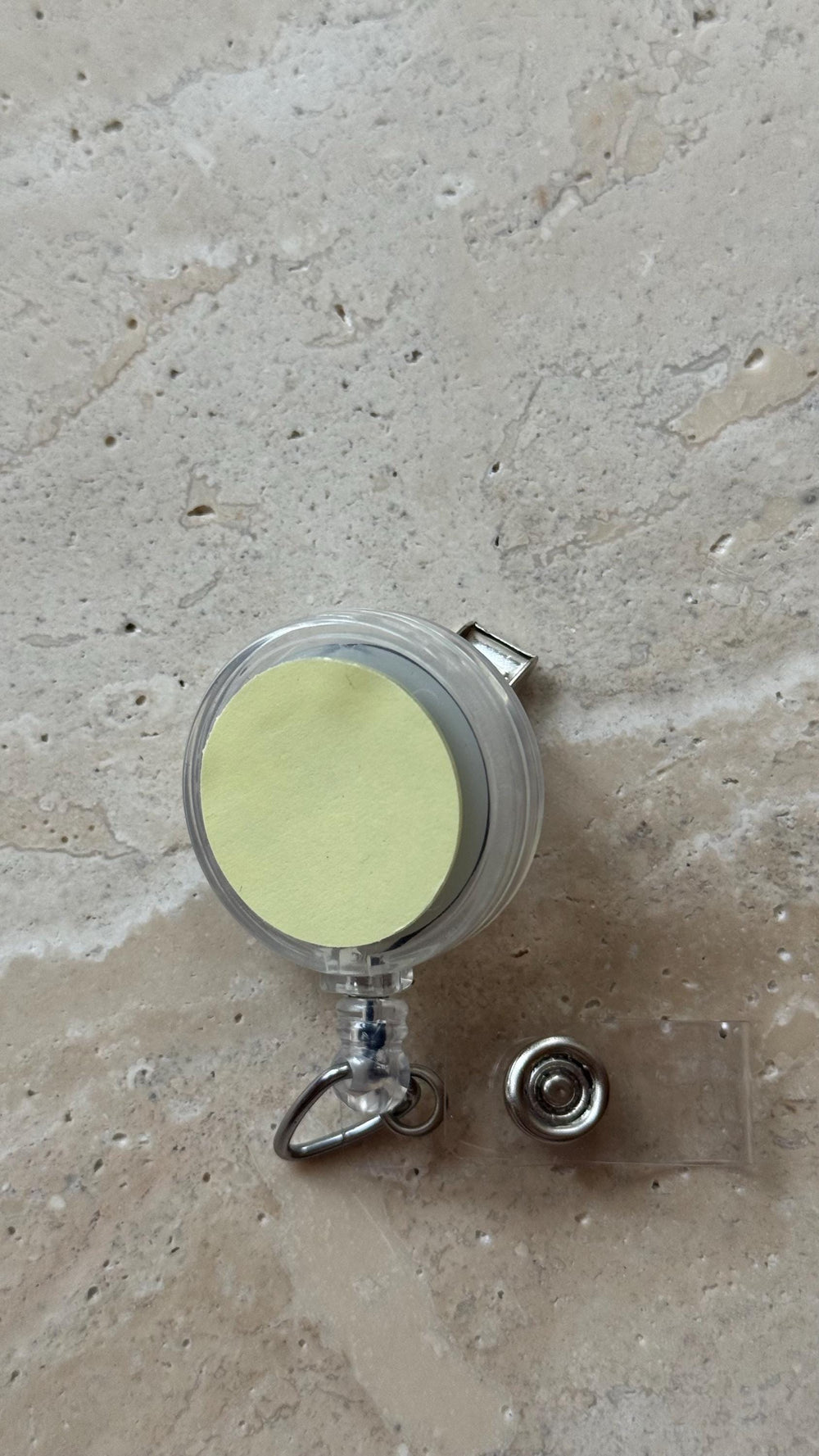 A clear plastic retractable badge reel with a silver metal clip and a clear plastic strap. The reel has a light yellow circular sticker on the front. The badge reel is designed to hold an  or other sm