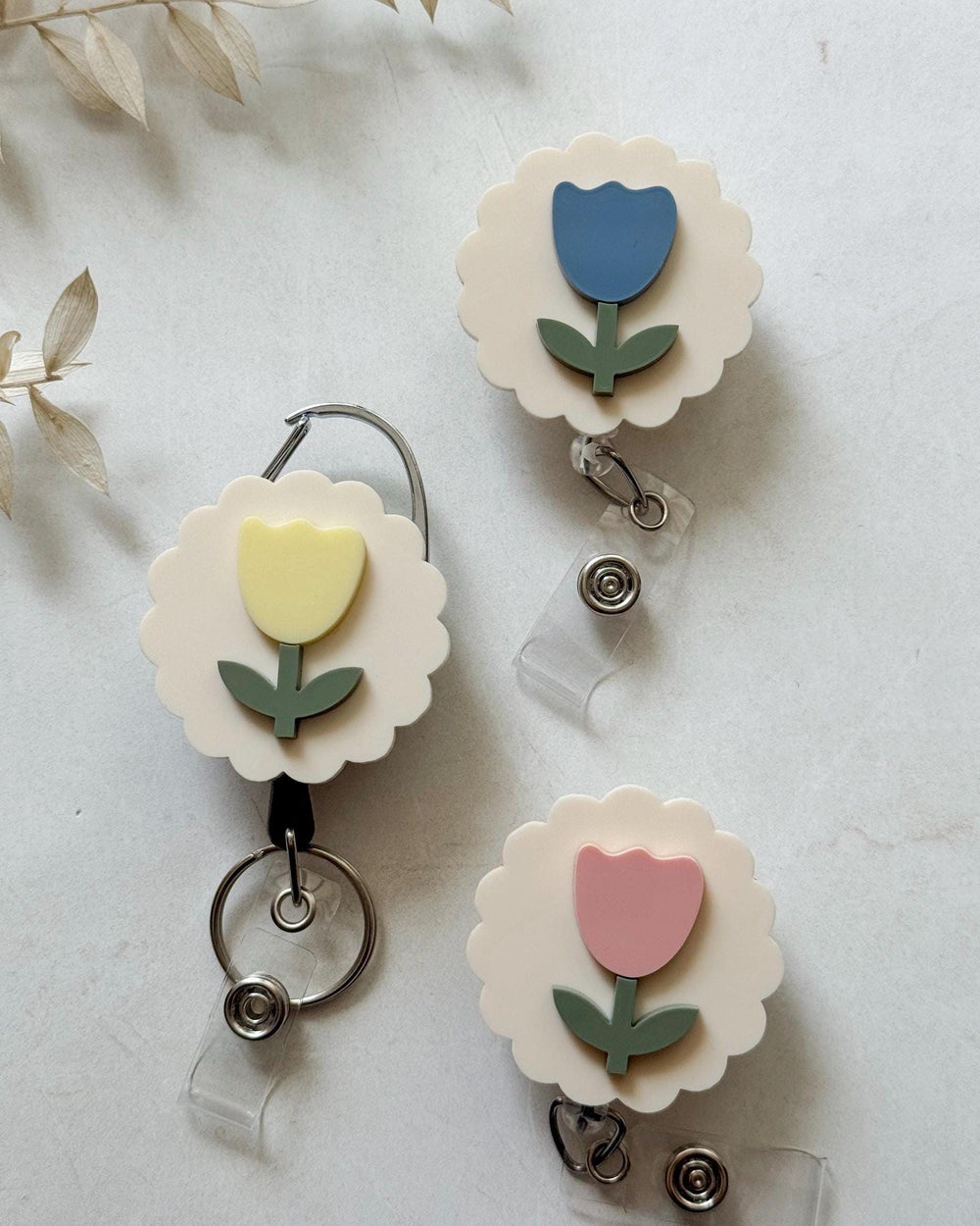 Three floral-shaped badge reels in pastel colors. Each reel features a flower design with a scalloped edge, a colored tulip, and a green stem. The reels are in yellow, blue, and pink.