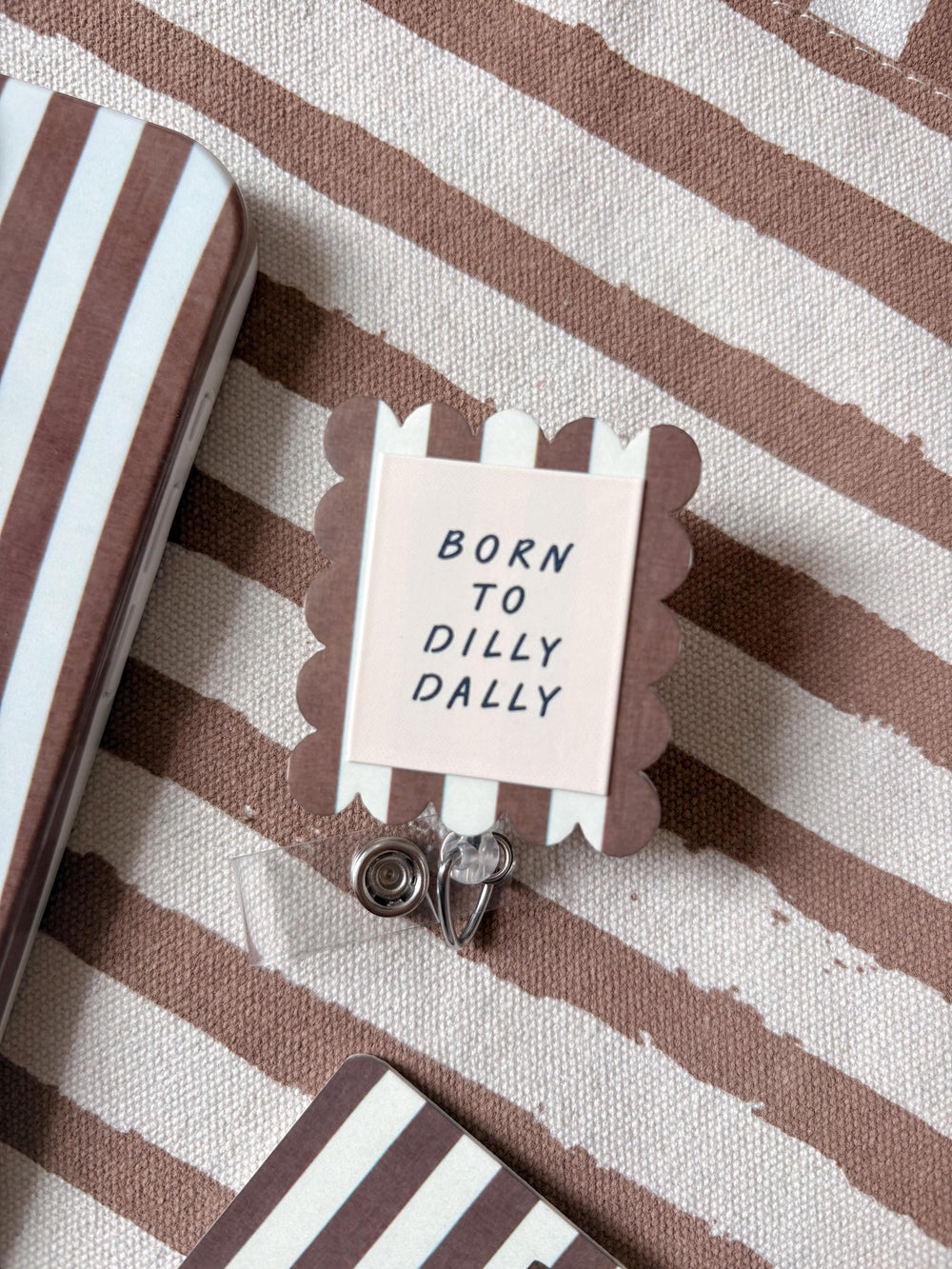 A square badge reel with scalloped edges featuring brown and white vertical stripes. The center is a light cream square with the black text BORN TO DILLY DALLY. A clear plastic strap with a metal snap