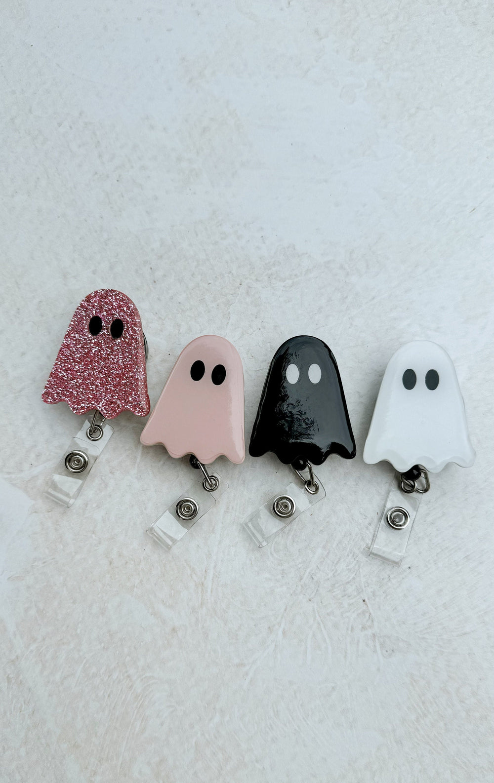 Four ghost-shaped retractable badge holders in pink glitter, light pink, black, and white. Each ghost has two black eyes and a clear plastic retractable reel with a metal clip.
