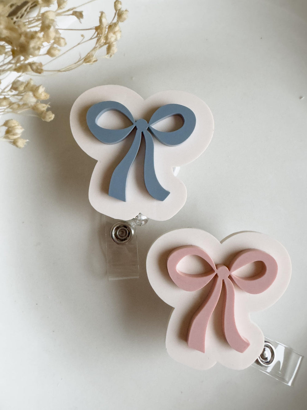Two badge reels with bow designs. One reel features a light blue bow, and the other has a pink bow. Both bows are on a white, cloud-shaped background. Each reel has a clear plastic clip and a retracta