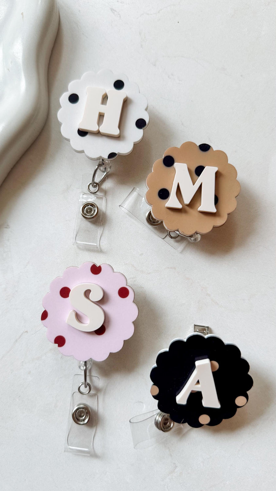 Four badge reels with scalloped edges and different colored backgrounds. Each reel features a white letter: H, M, S, and A. The reels have polka dots and a clear plastic strap with a metal clip.