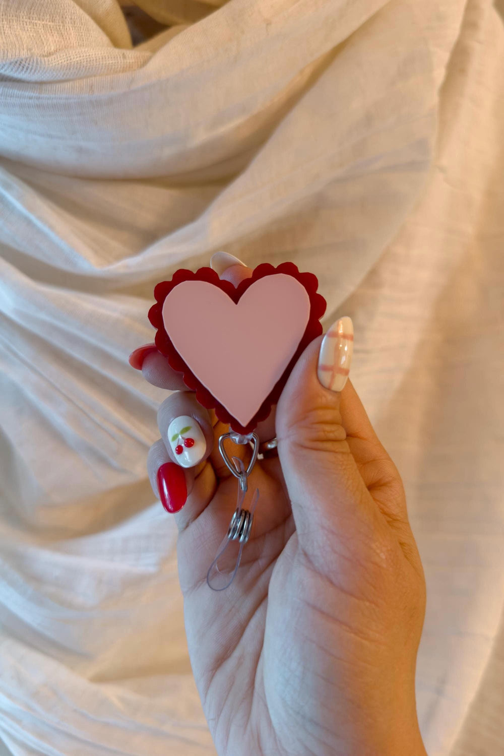 A heart-shaped badge holder with a light pink center and a scalloped red border. The badge holder is held by a silver-colored metal clip. The background is a white fabric.