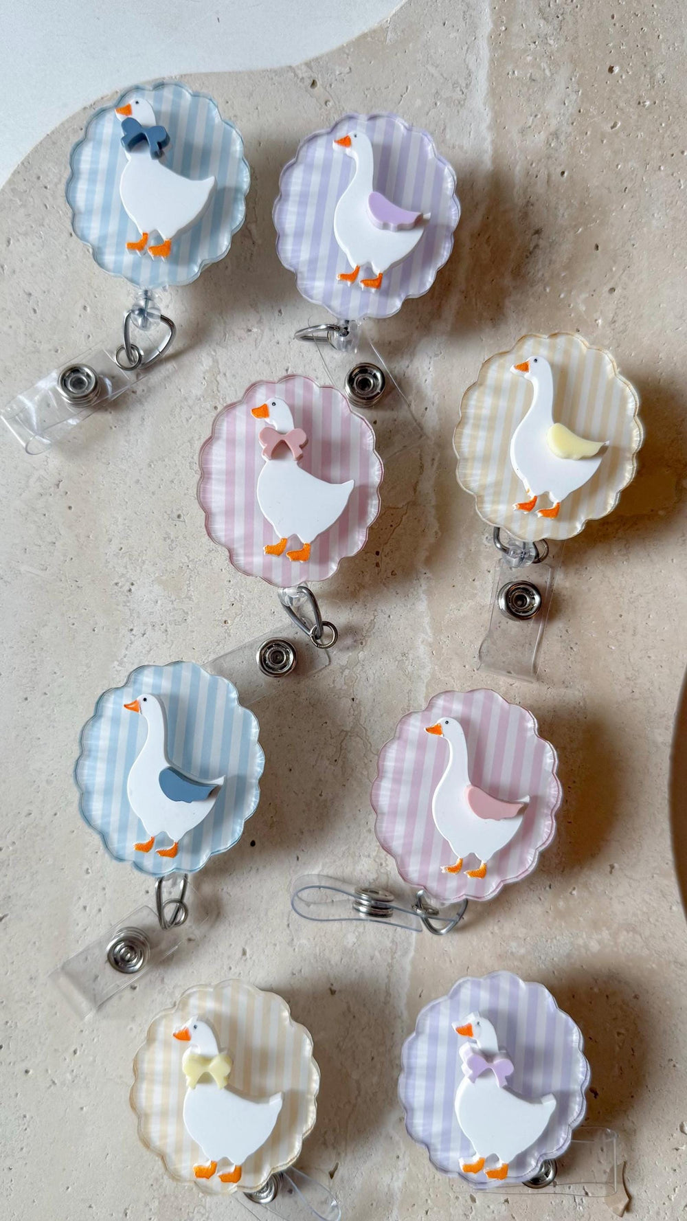 A collection of badge reels featuring a white goose design with orange feet and a bow. The reels have a scalloped edge and a striped background in various pastel colors, including blue, pink, and yell