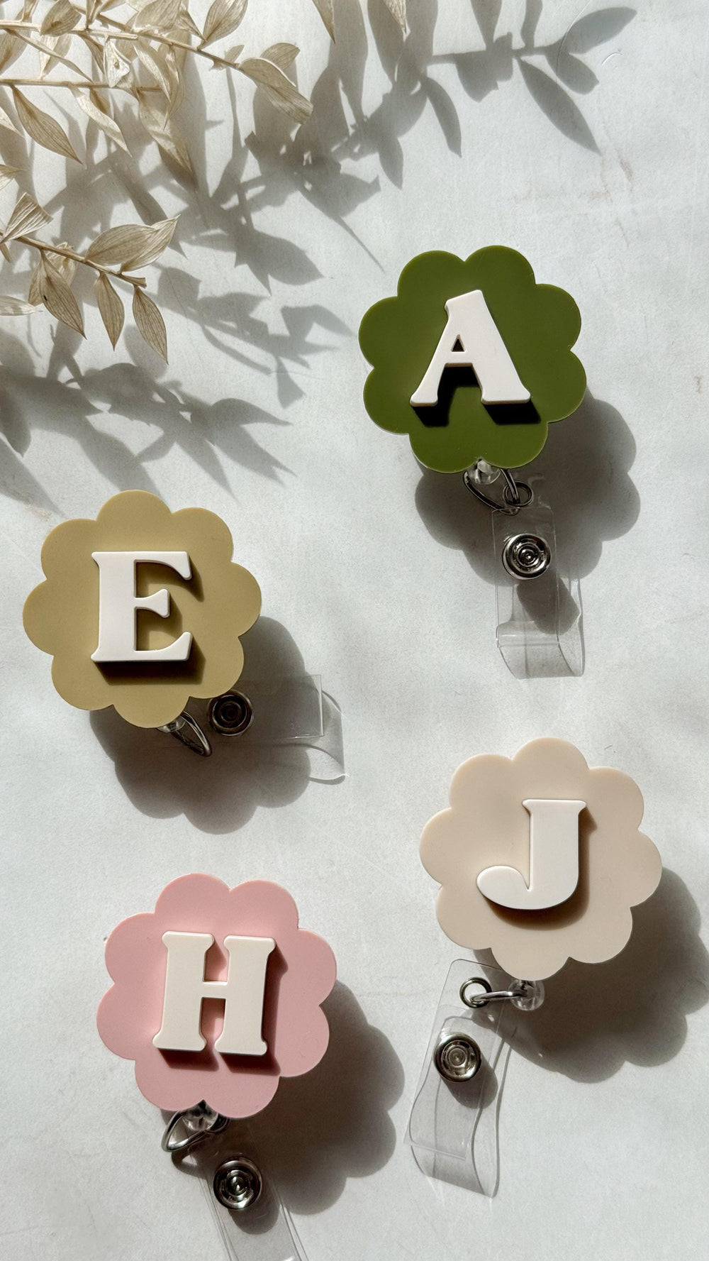 Four floral-shaped badge reels with the letters A, E, H, and J. The letters are white, and the flowers are in shades of green, tan, and pink. Each reel has a clear plastic retractable cord and a metal