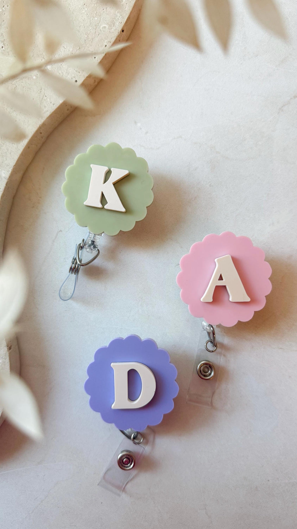 Three personalized badge reels with scalloped edges. The reels are in pastel colors: green, pink, and purple. Each reel has a white letter: K, A, and D. The reels have a clear plastic strap and a meta
