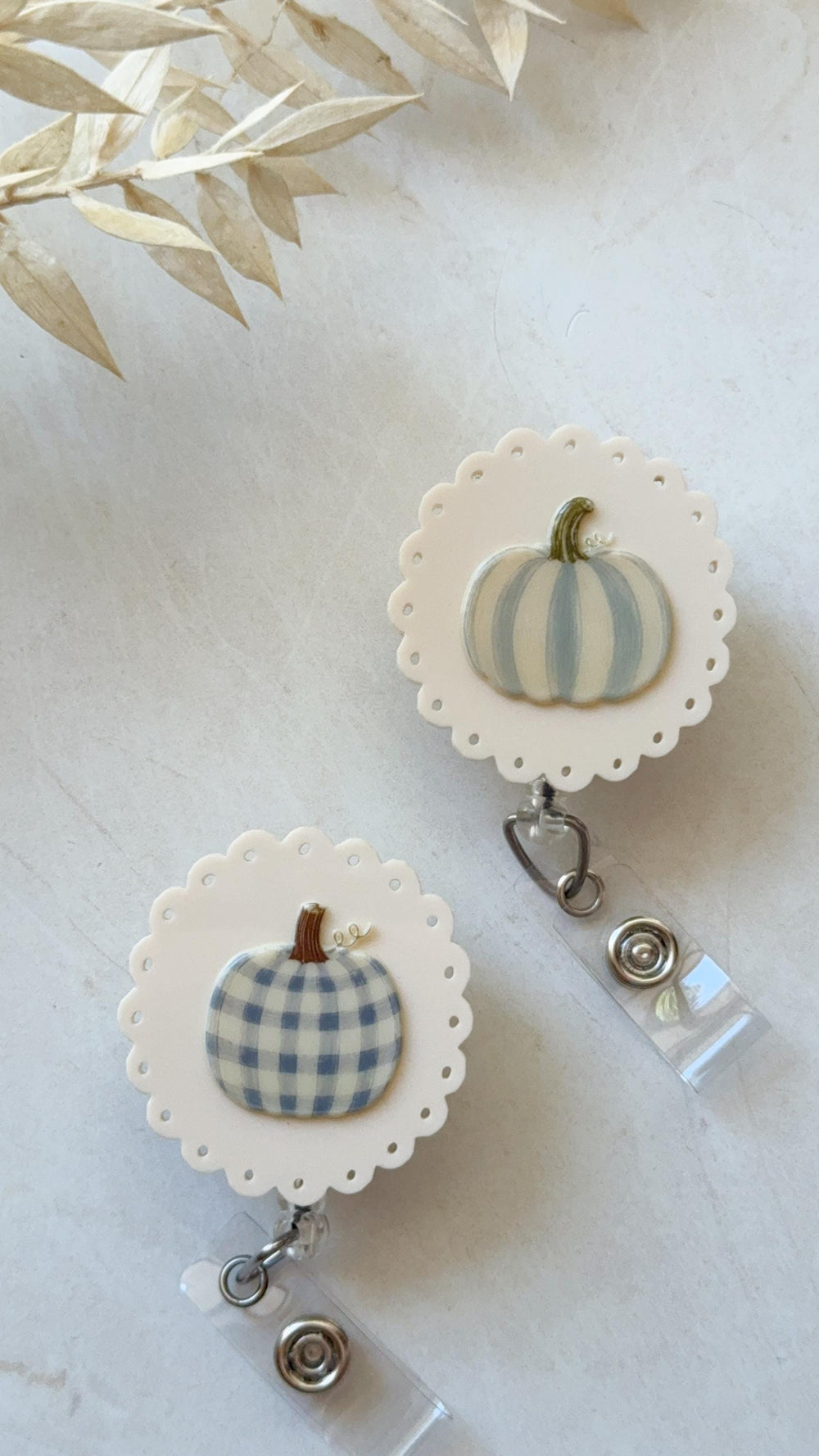 Two round badge reels with scalloped edges and white backgrounds. One features a light blue and white striped pumpkin, while the other displays a light blue and white gingham patterned pumpkin. Both i