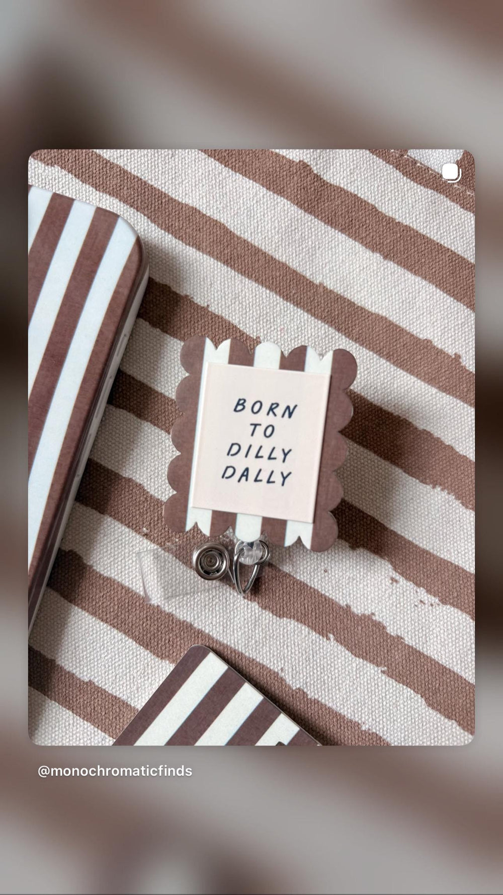 A square badge reel with a scalloped edge featuring brown and white vertical stripes. The center has a light pink square with the black text BORN TO DILLY DALLY. A clear plastic strap with a metal sna