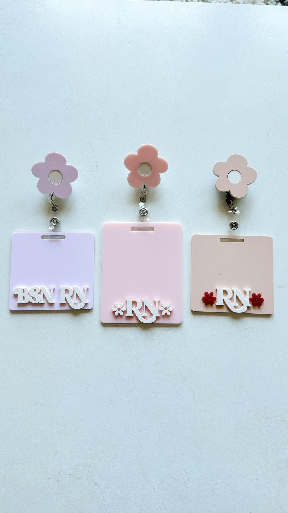 Three pink and white flower-shaped badge holders with retractable reels. Each badge holder has a different design: one says "BSN ", one says "**" with a flower on each side, and one says "" with a red