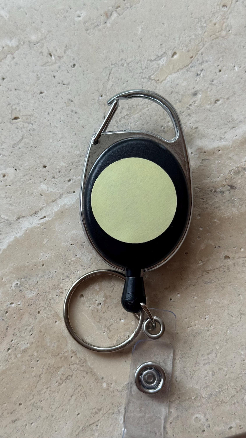 A black retractable badge reel with a silver carabiner clip and key ring. The reel has a round, light yellow sticker in the center. A clear plastic strap with a snap closure is attached.
