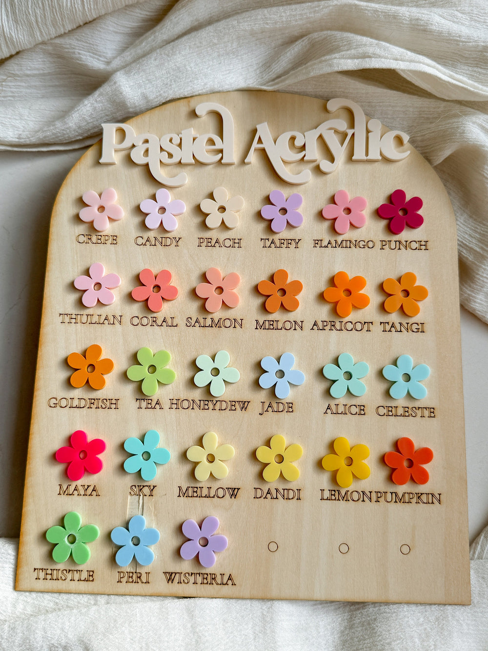 A wooden board with the text "Pastel Acrylic" at the top. The board is divided into rows with different colored flower-shaped acrylic pieces. Each piece has a name written below it, such as "Crepe", "