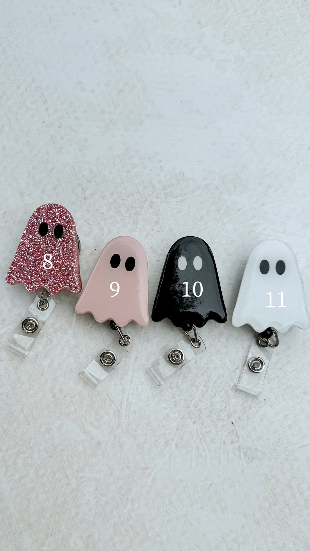 Four ghost-shaped badge reels in pink glitter, light pink, black, and white. Each ghost has black eyes and a number from 8 to 11. The reels have a clear plastic strap and a metal clip.