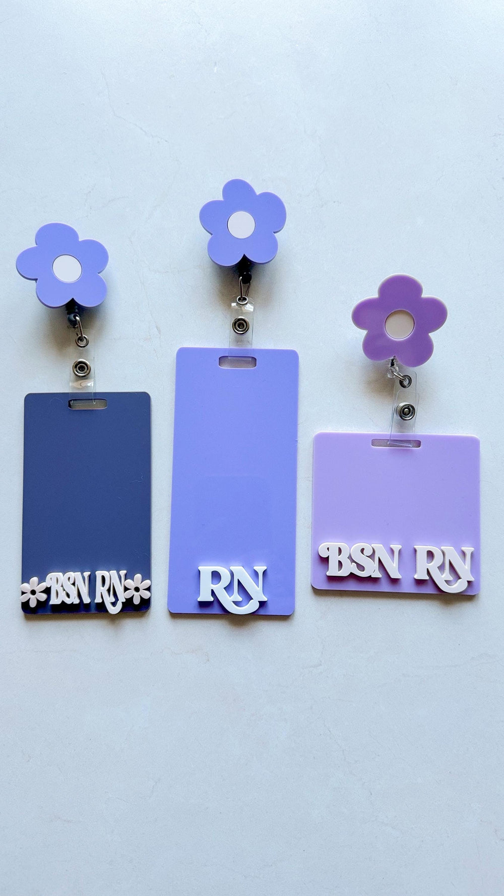 Three purple and white flower badge reels with different text on the badge holders. The left badge holder says "BSN " with a flower on each side. The middle badge holder says "" and the right badge ho