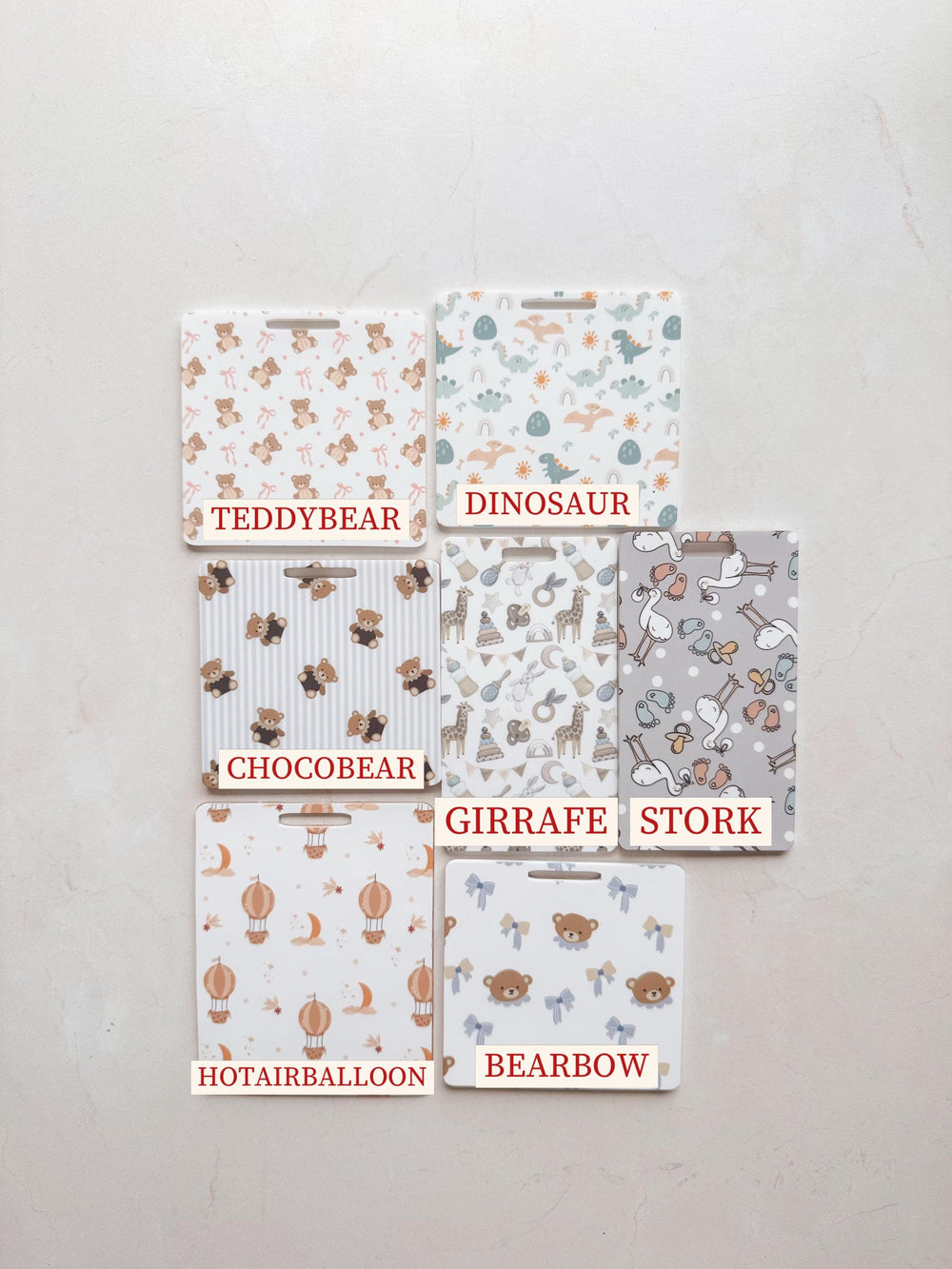 Six rectangular baby-themed placemats with different patterns. Designs include teddy bears, dinosaurs, giraffes, storks, hot air balloons, and bear bows. Each placemat has a white background with a un
