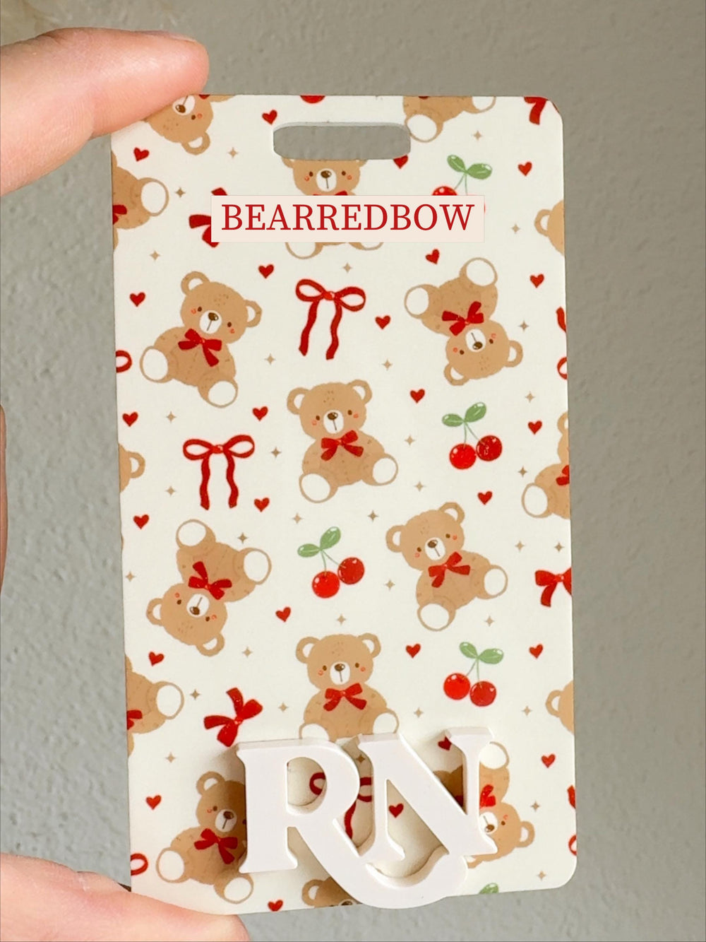 A rectangular badge with a repeating pattern of brown teddy bears, red bows, cherries, and hearts on a cream background. The text "BEARREDBOW" is at the top, and the letters "" are at the bottom. The 