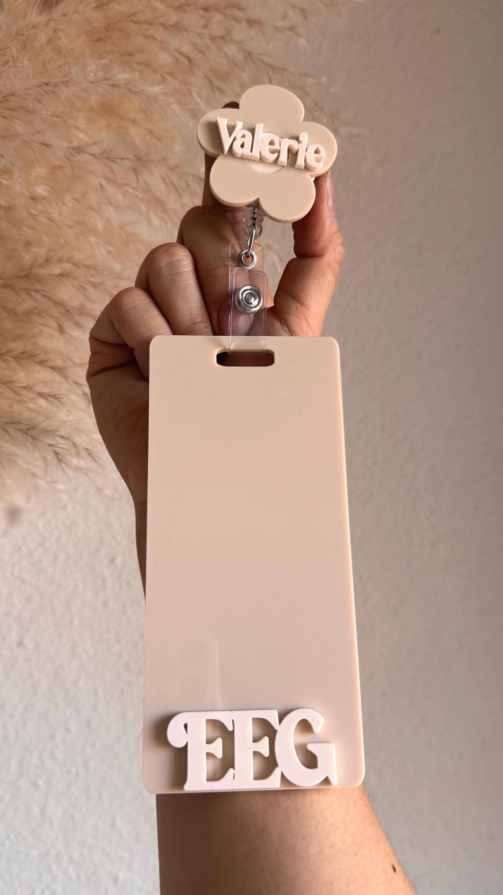 A beige plastic badge holder with a flower-shaped retractable badge reel. The badge holder has a slot for a name tag and the text "EEG" is printed on the bottom. The badge reel has the name "Valerie" 
