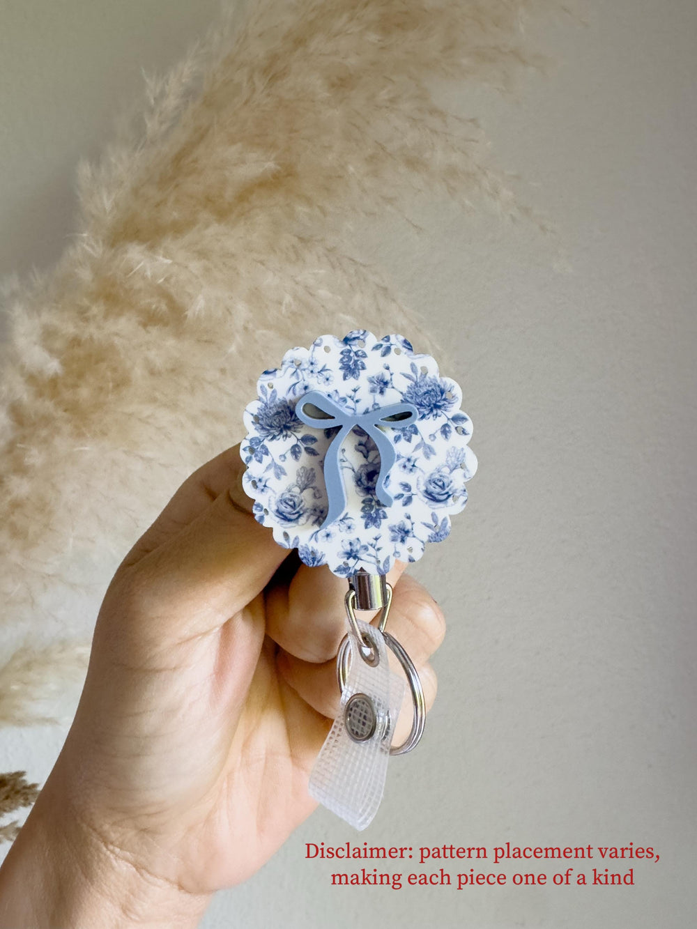 A floral badge reel with a blue bow accent. The reel features a white background with blue floral patterns. The reel is attached to a silver metal clip and a clear plastic strap. The text "Disclaimer: