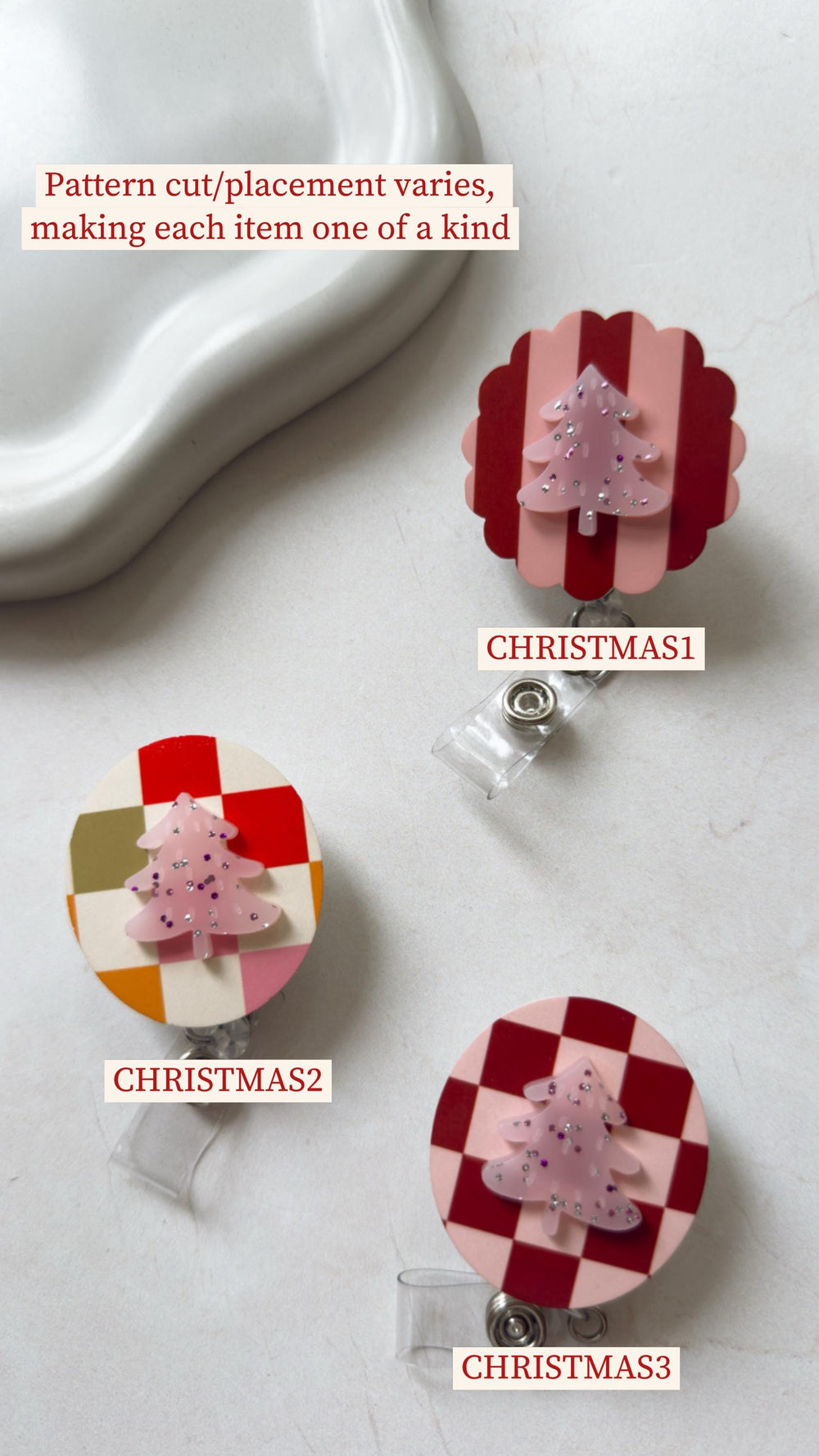 Three Christmas-themed badge reels with different designs. One has a red and pink striped background, another has a checkered pattern, and the third has a color block design. Each reel features a pink