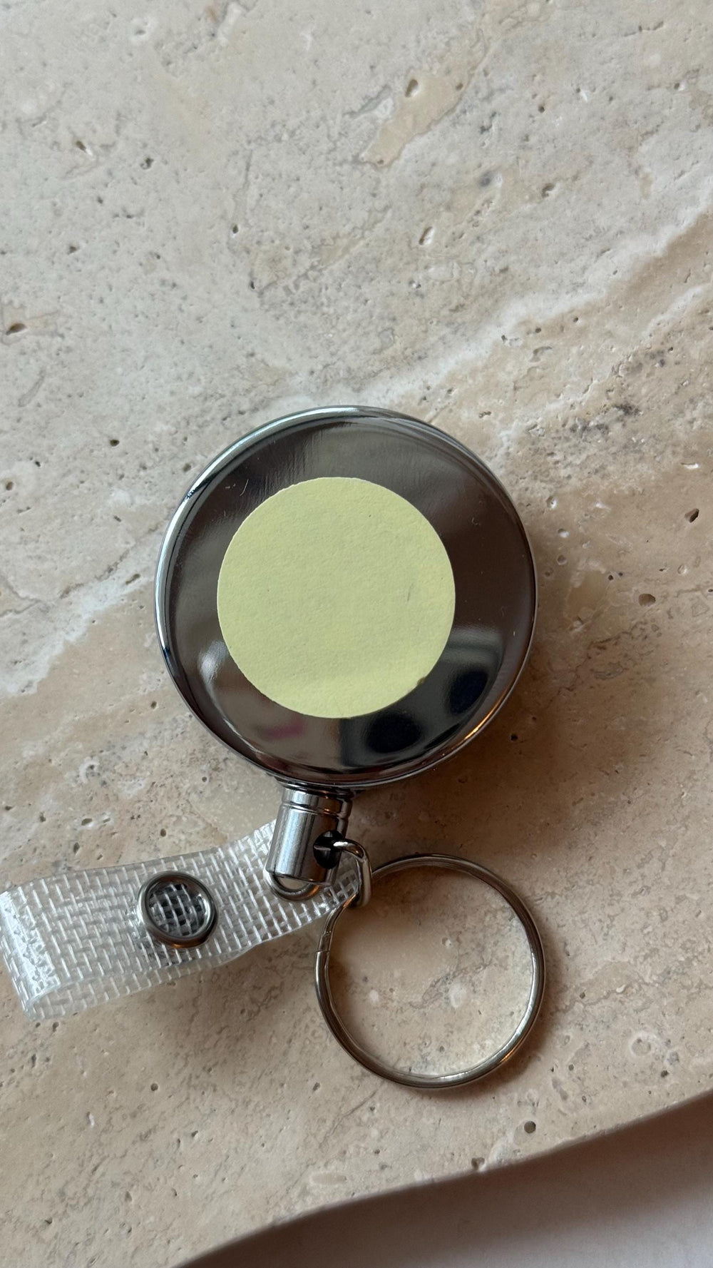 A silver retractable badge reel with a clear plastic strap and a key ring. The reel has a yellow circular sticker in the center. The background is a light brown, textured surface.