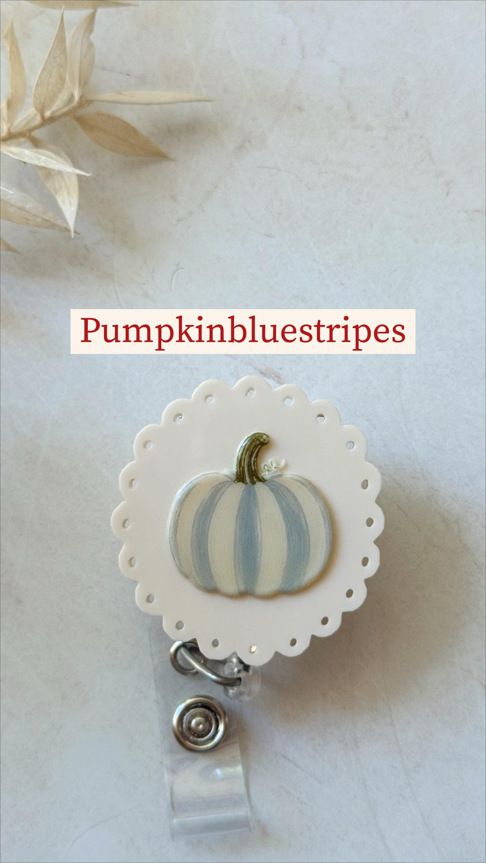 A round white badge reel with a scalloped edge featuring a painted pumpkin with light blue and white vertical stripes and a green stem. The text Pumpkinbluestripes is displayed above the badge reel, w