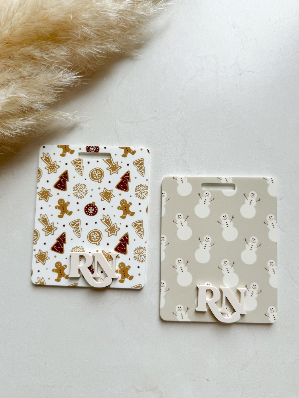 Two white plastic name tags with a Christmas theme. One tag has a gingerbread man and Christmas tree pattern, the other has a snowman pattern. Both tags have the letters "" in white.