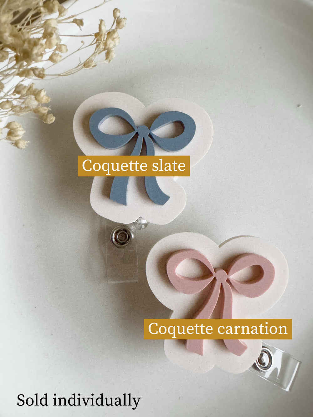 Two badge reels with bow designs. One reel has a slate blue bow, the other has a carnation pink bow. Each bow is on a white, cloud-shaped background. The text "Coquette slate" and "Coquette carnation"