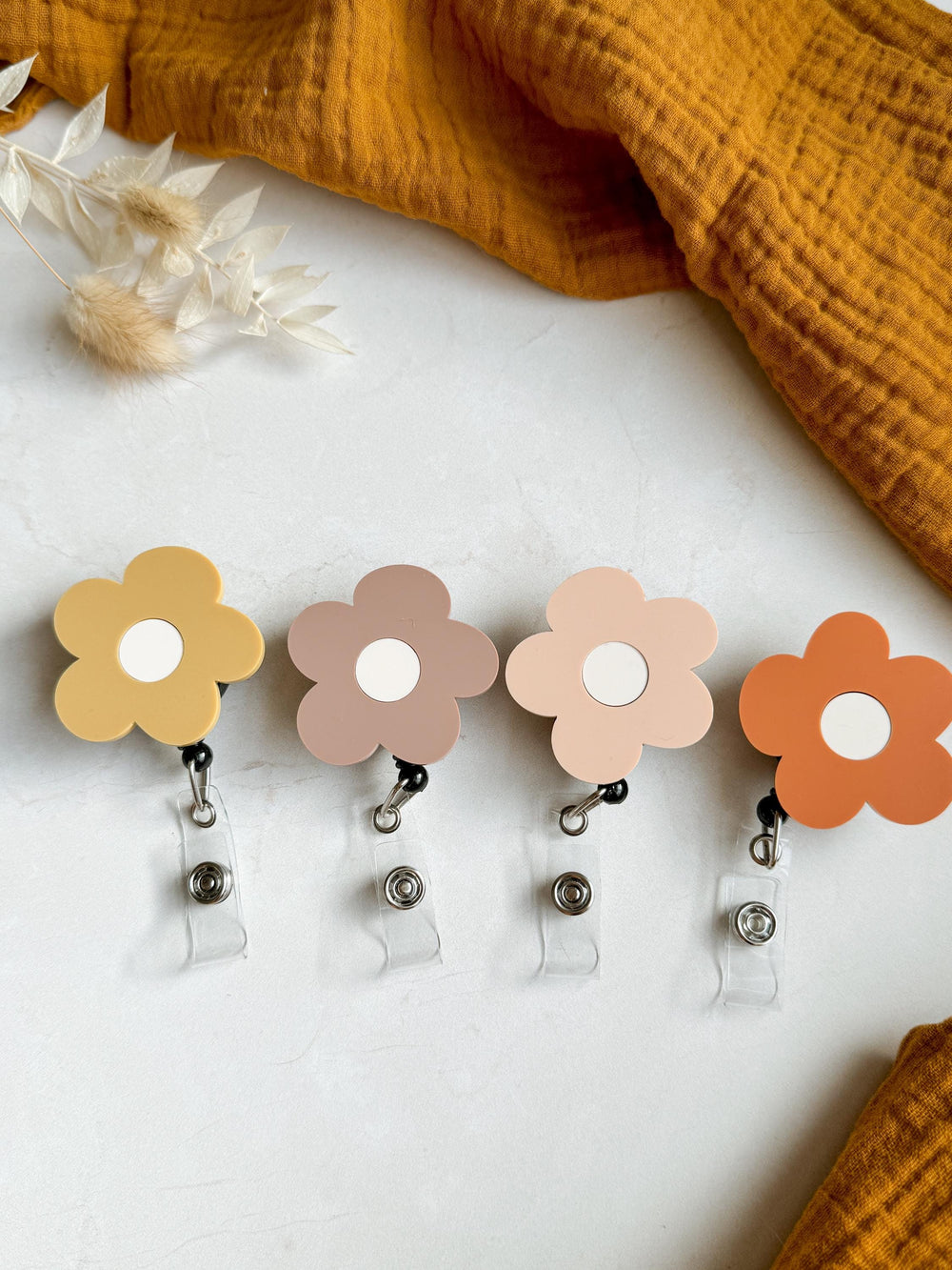 Four flower-shaped badge holders with a white center and a black clip. The colors of the flowers are yellow, brown, pink, and orange.