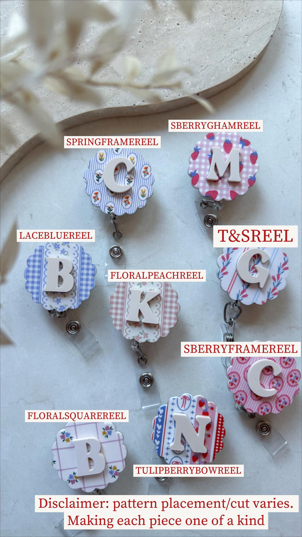 A collection of decorative badge reels with various patterns and initial letters. Designs include floral, gingham, and strawberry patterns. Each reel has a clear plastic strap and a metal clip. The te