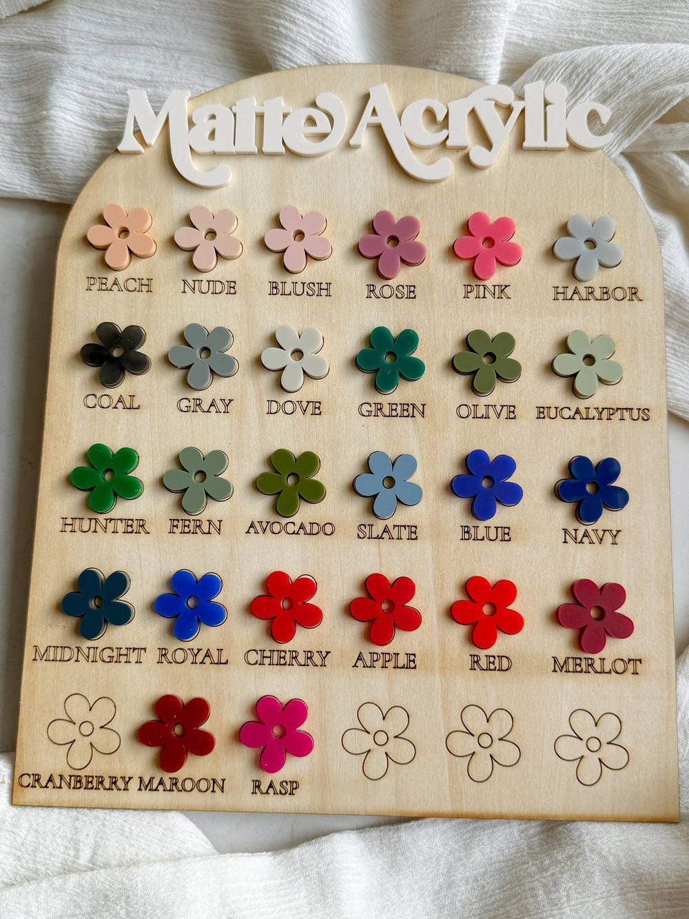 A wooden display board with the words "Matte Acrylic" at the top. The board is divided into 18 sections, each containing a small flower-shaped piece of acrylic in a different color. The colors are lab