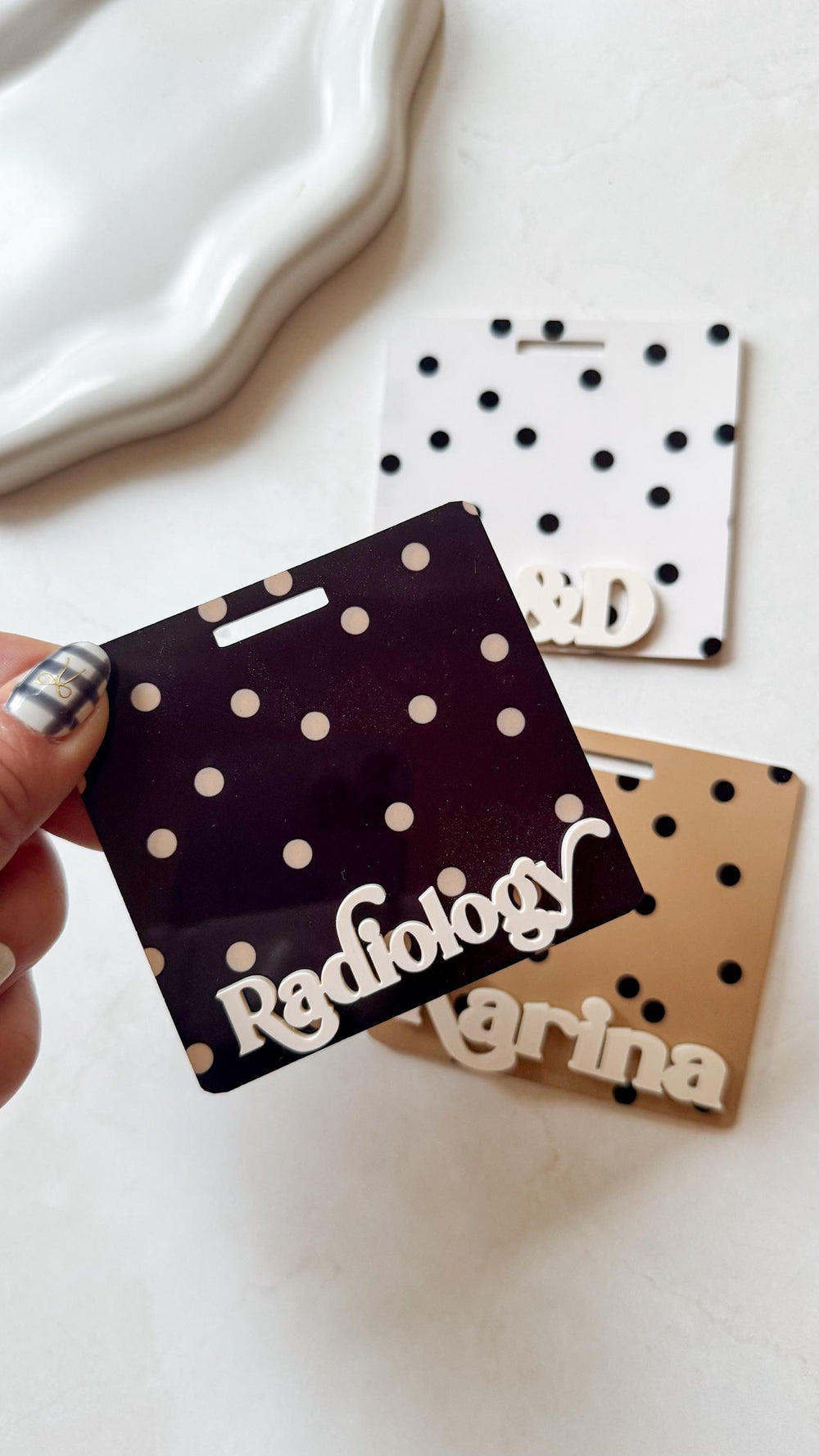 Three name badges with a polka dot pattern. One is dark purple with the word "" in white. Another is white with black dots and the letters "S&D". The third is tan with the name "Marina".