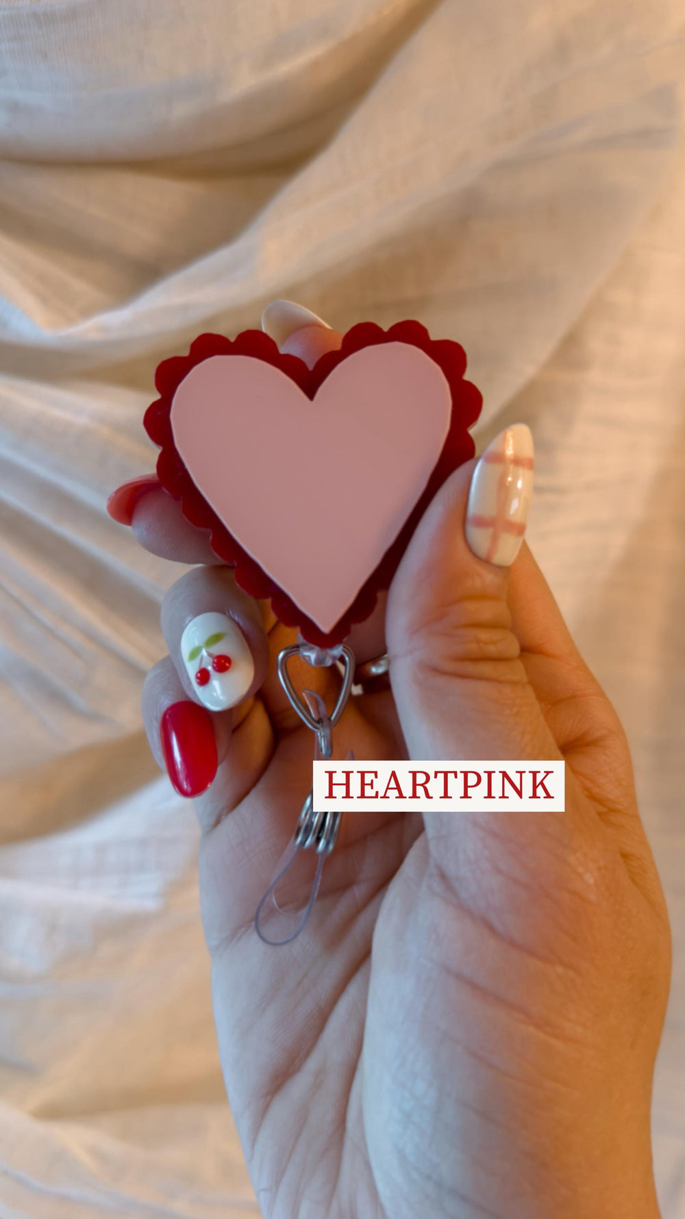 A pink heart-shaped badge with a red scalloped border. The badge has a silver-colored clip and the text "HEARTPINK" in a white rectangle. The image shows a close-up of the badge.