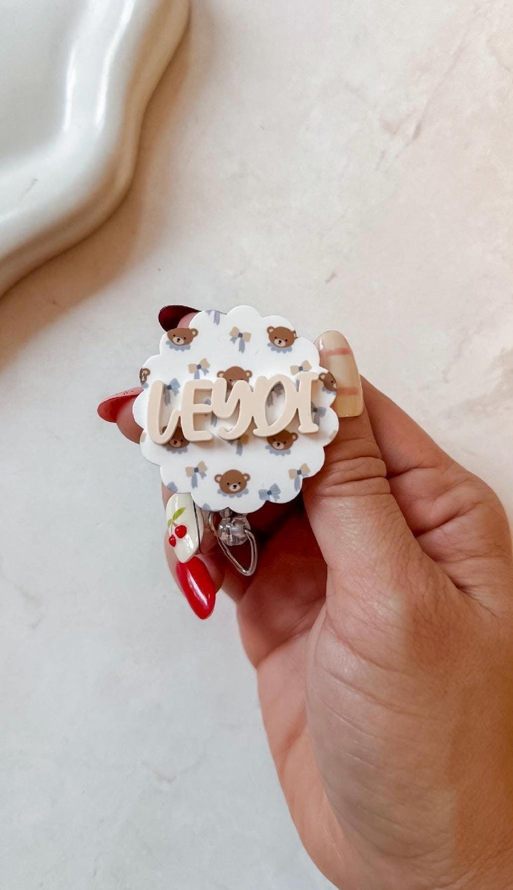 A white badge with a scalloped edge, featuring a pattern of teddy bears and blue bows. The name "LEYDI" is displayed in a light brown, 3D font across the center. A silver badge reel is attached.
