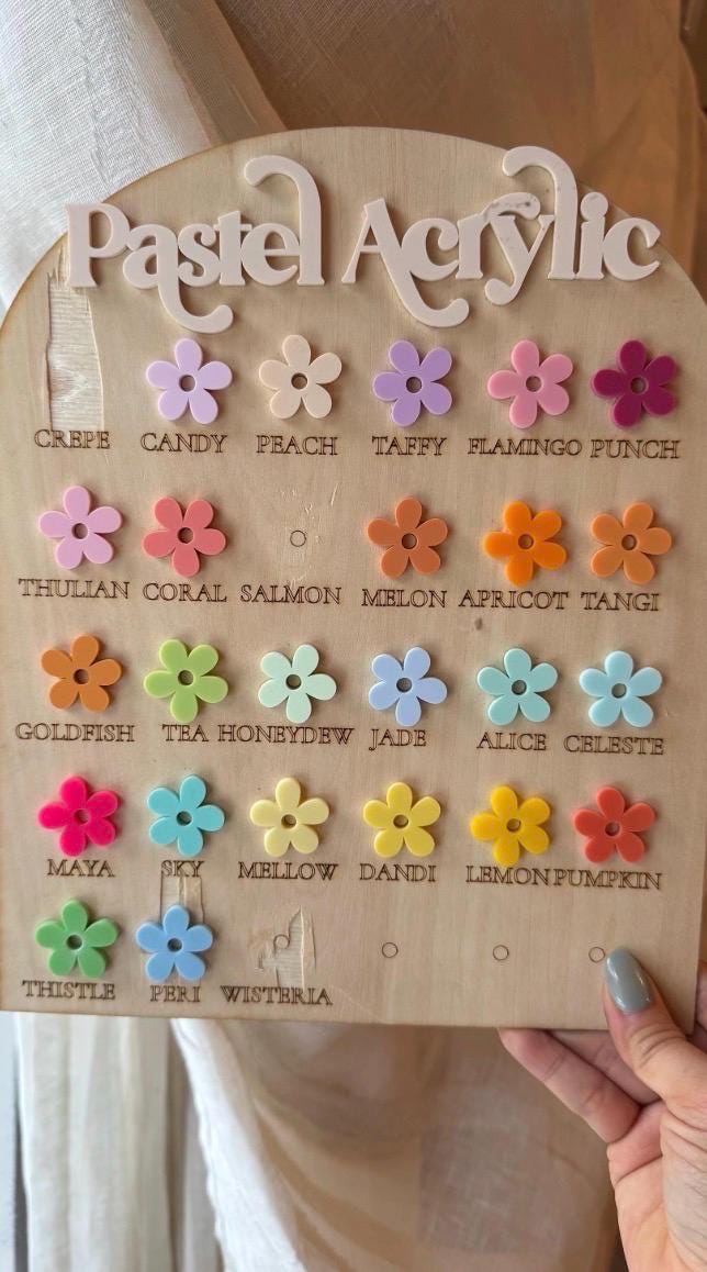 A wooden display board with the words "Pastel Acrylic" and colorful flower-shaped acrylic pieces. The flowers are in various pastel shades, including pink, orange, yellow, blue, and green. Each flower