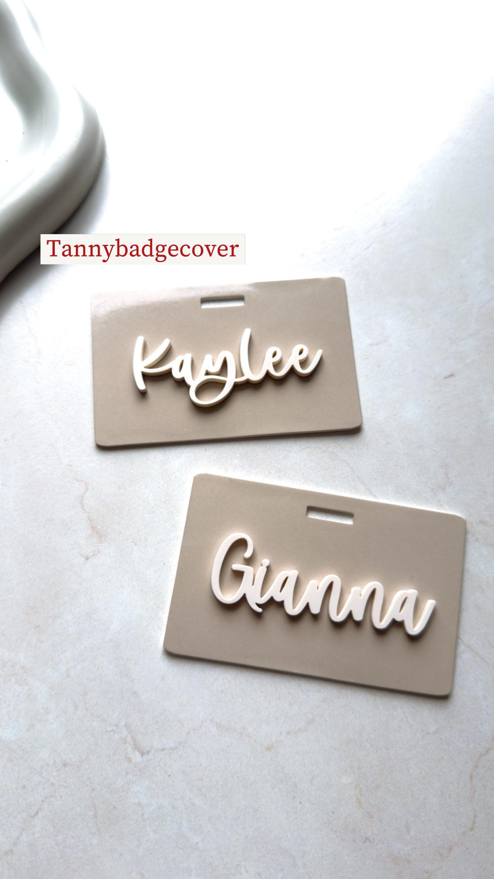 Two beige name badges with white cursive lettering. The top badge reads "Kaylee" and the bottom badge reads "Gianna". The text "Tannybadgecover" is in the top left corner.