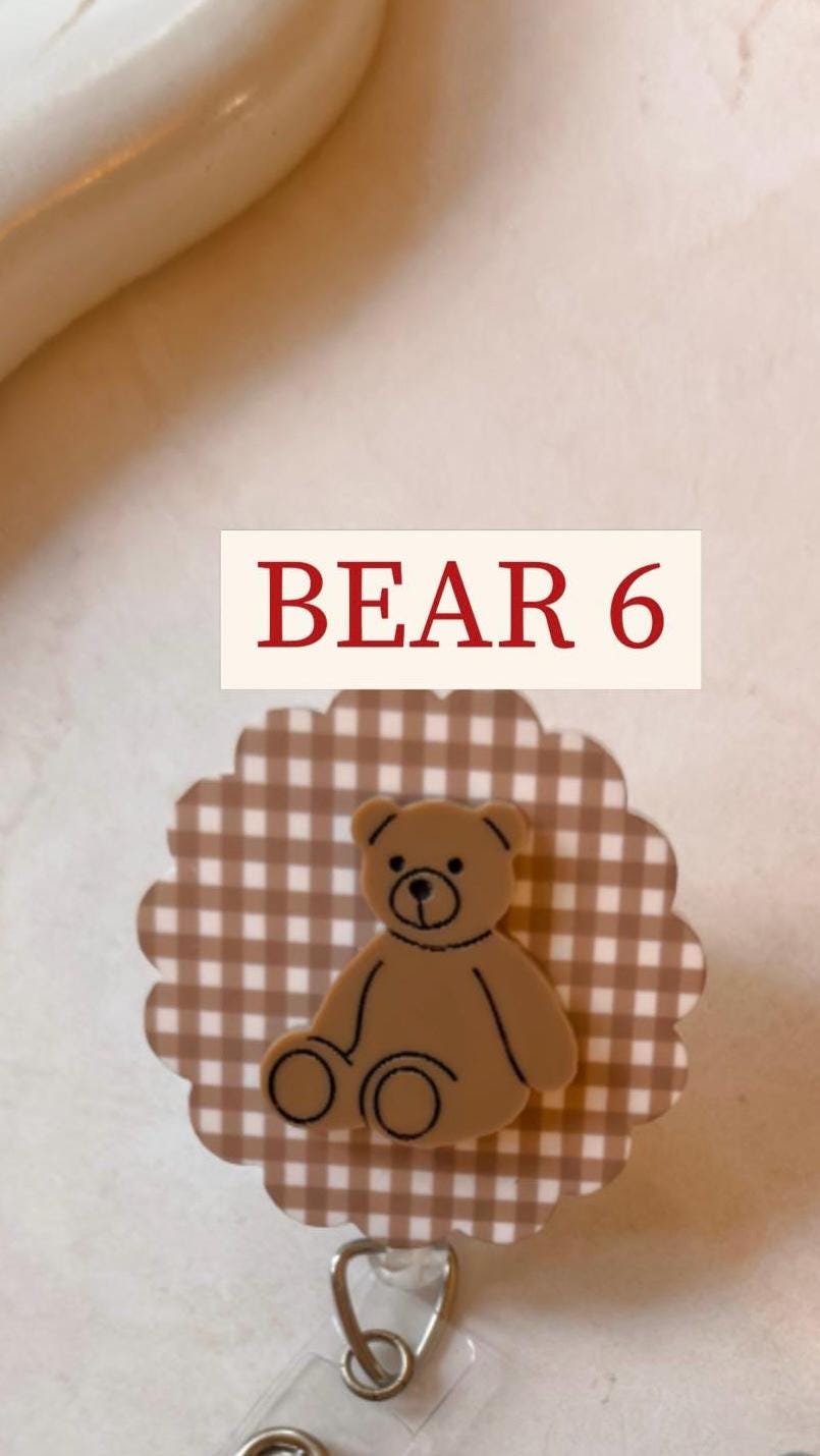 A brown bear-shaped badge reel with the text "BEAR 6" in red. The bear is on a brown and white checkered background with a scalloped edge. The reel has a silver-colored clip.