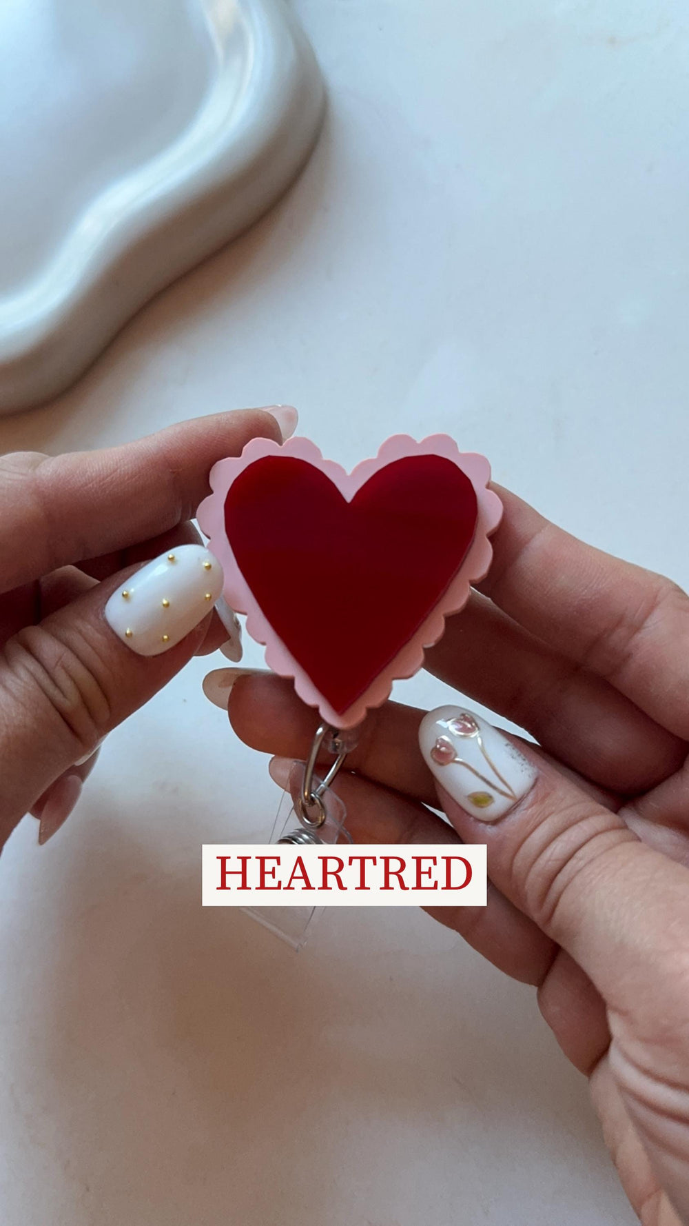 A red heart-shaped badge reel with a scalloped pink border. The text "HEARTRED" is displayed in a white rectangle. The badge reel is held in front of a white background.