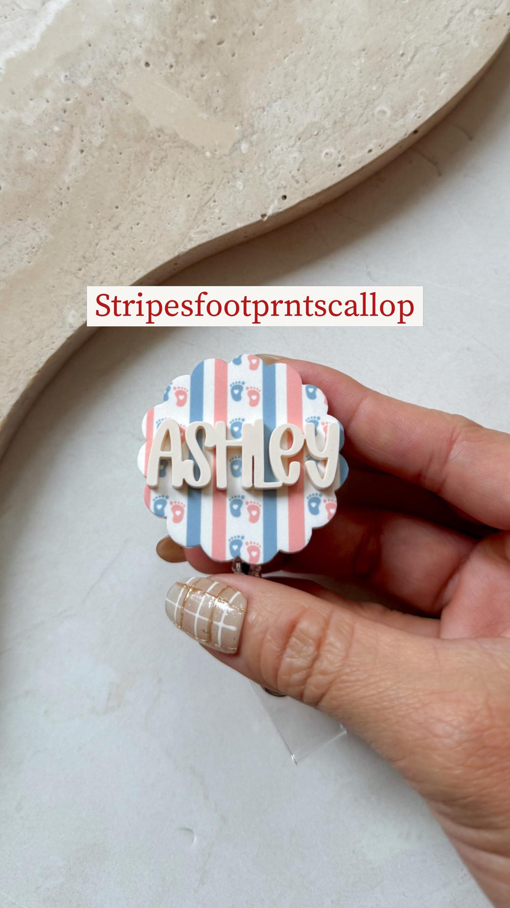 A badge with a scalloped edge, featuring a striped pattern in blue and pink with tiny footprints. The name "Ashley" is displayed in white, three-dimensional letters. The text "Stripesfootprntscallop" 