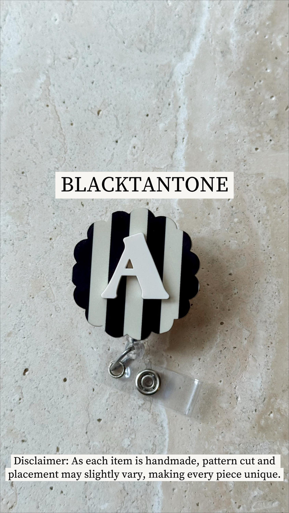 A badge reel with a black and white striped design and a white letter "A". The reel has a scalloped edge and a clear plastic clip. The text "BLACKTANTONE" is at the top.