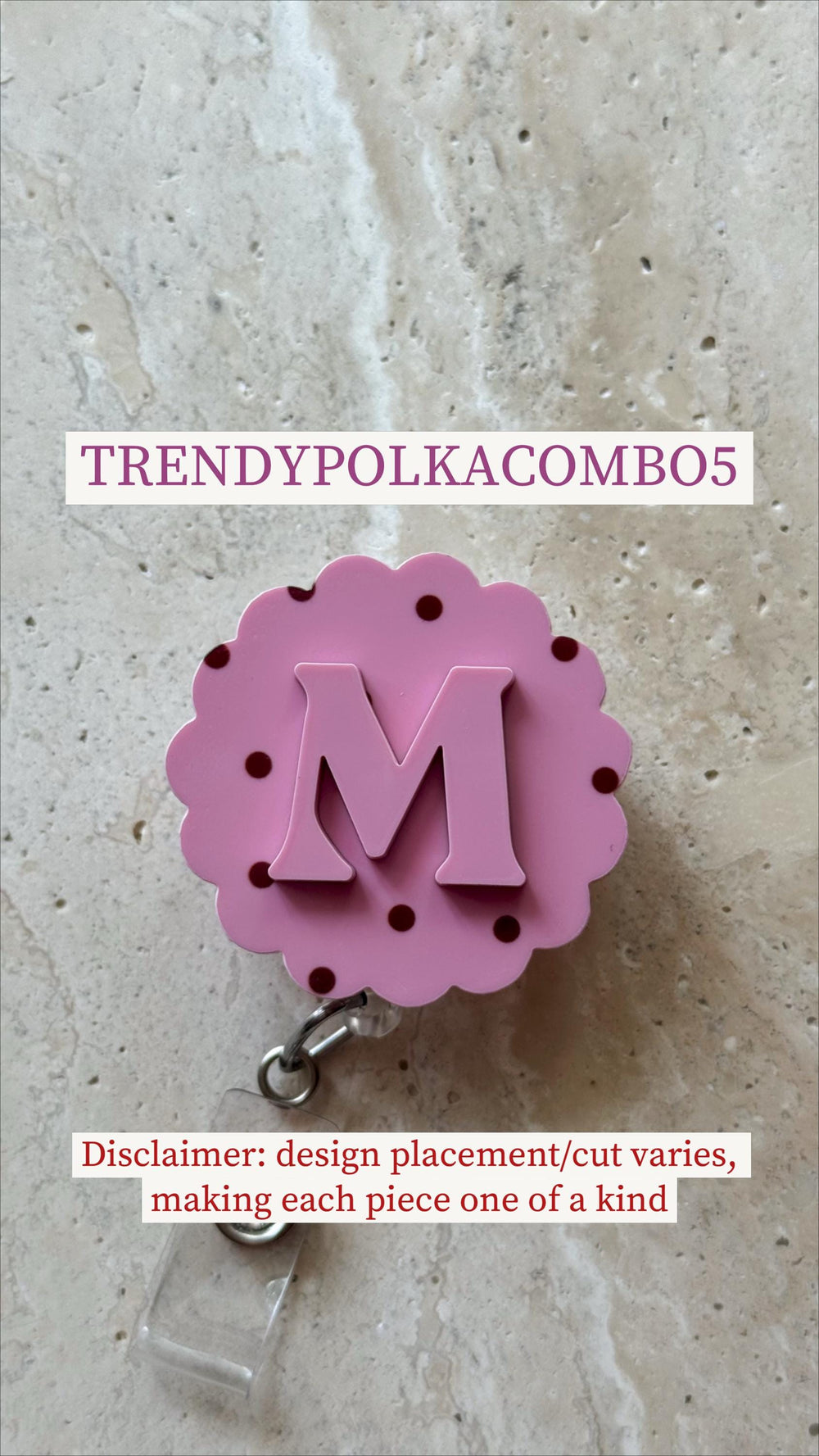 A pink badge reel with a scalloped edge and the letter "M" in the center. The reel has dark pink polka dots and the text "TRENDYPOLKACOMBO5". The reel is attached to a clear plastic clip.